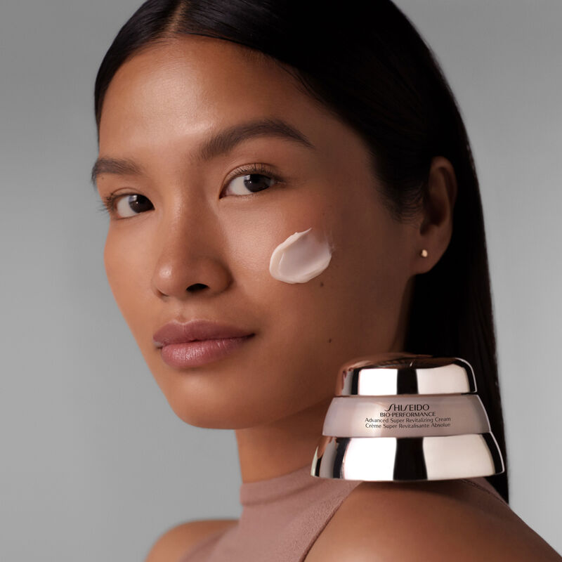 "A model with the Shiseido Bio-Performance Advanced Super Revitalizing Cream applied on her cheek as she looks toward the camera,showcasing the rich-textured cream. The product's jar is also perched on her left shoulder. "