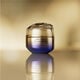 Un frasco redondo, closed gold-and-purple-colored jar of Shiseido Vital Perfection Uplifting and Firming Advanced Cream. The jar is shown against a blurred gold background.