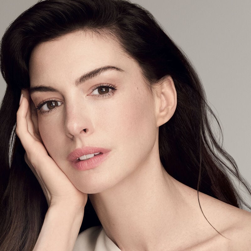The actress Anne Hathaway holding a closed jar of Shiseido Vital Perfection Uplifting and Firming Advanced Cream against a white background. The closed jar features the Shiseido logo, el nombre del producto, y las palabras "ReNeuraRED Technology."