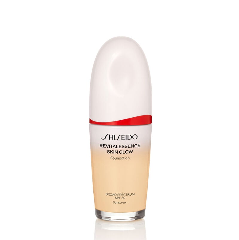 An oval-shaped bottle of the Shiseido Revitalessence Skin Glow Foundation SPF 30 in shade 130 Opal. The label on the bottle is white with a red accent. El texto en la etiqueta muestra el título del producto, y su protección solar.